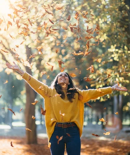 Casual joyful woman having fun throwing leaves in autumn at city park.